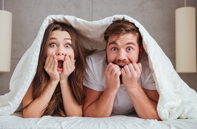 Surprised young loving couple looking at camera while lying in bed under blanket and holding their face with hands.