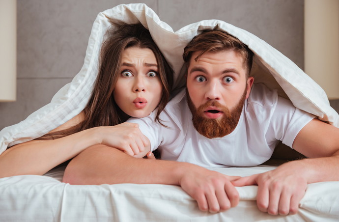 Shocked young loving couple looking at camera while lying in bed under blanket