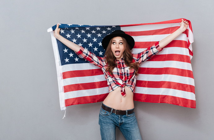 Portrait of a cheerful excited young woman holding USA flag and looking away isolated over gray background